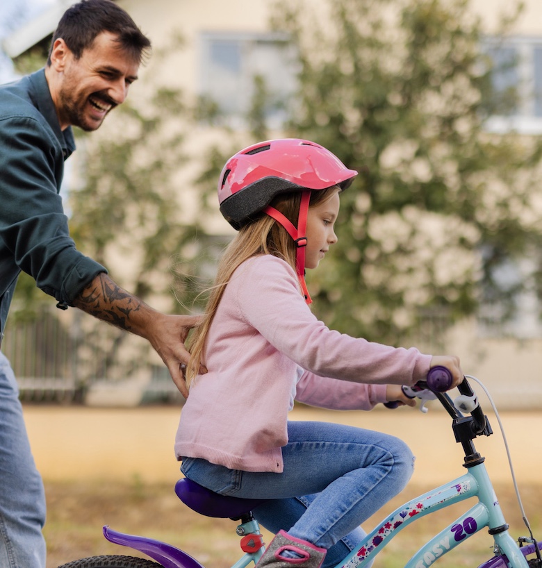 Father Helps Little Daughter to Ride Bicycle on the Sidewalk in the City Suburbs