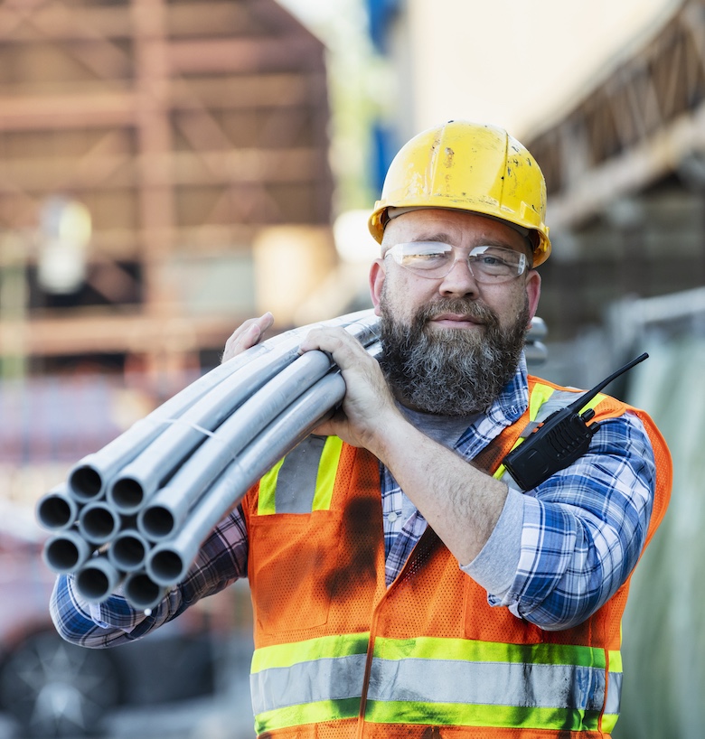 Construction worker wearing plastic protective gear carrying pipes on shoulder